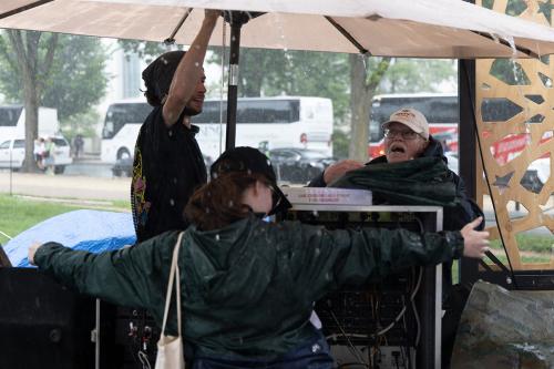 Three people gather under an umbrella while it rains. One has their back turned and arms outstretched, while another is adjusting the umbrella, and the third watches.