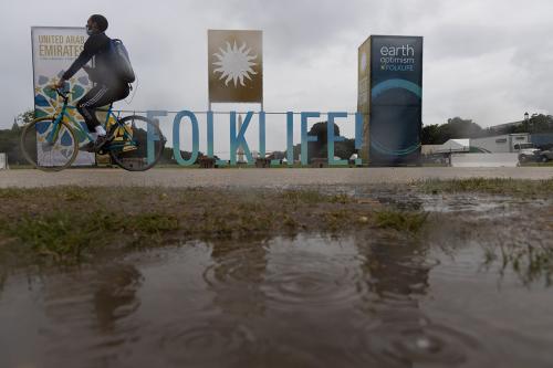 An outdoor marquee that reads “22 FOLKLIFE” in large, block letters under a gloomy sky. In the foreground, a biker goes through puddles on a brown gravel road.