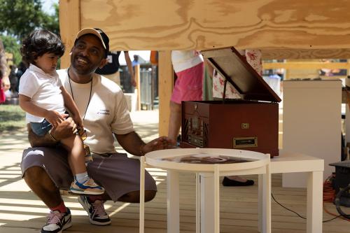 A man kneels outdoors, holding a child on one knee, next to a record player.