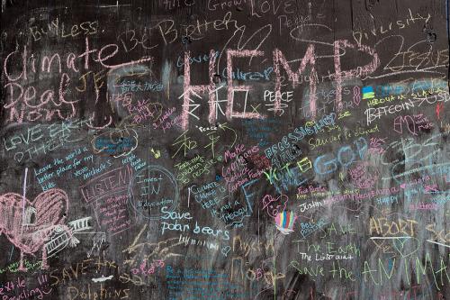 A blackboard with many messages written in pastel-colored chalk: Climate Deal Now, Love Each Other, LISTEN to Indigenous Americans!!! Save polar bears, HEMP, Teach my children to recycle, Buy Less.