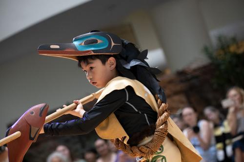 A child wearing a bird-like wooden headpiece holding a stick with a wooden fish on it.