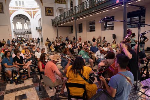 In a grand hall, a circle of musicians play string instruments for a seated crowd.