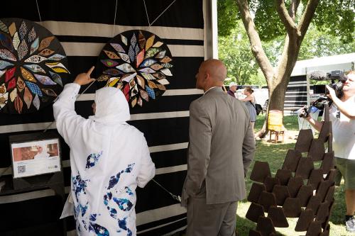 A woman in white dress and headscarf and a man in tan suit look at a piece of artwork inside a tent, while someone standing to the side films with a video camera. In the foreground, a metallic sculpture.
