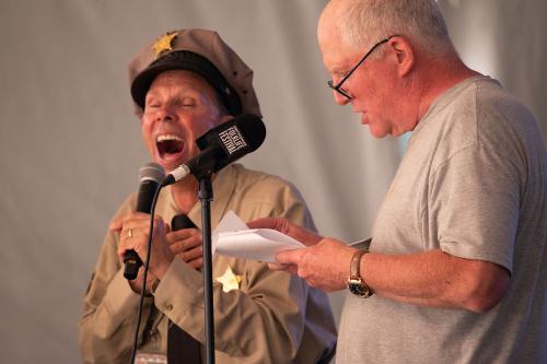 A man in a police officer uniform laughs hysterically as a man reads from a paper into a microphone.
