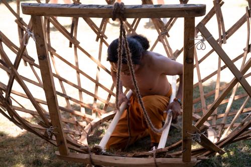 A man in yellow pants and no shirt kneels in the grass, holding two parallel poles at the rooftop opening of an upside-down wooden yurt structure.