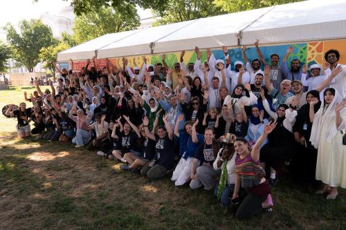 A group of dozens of people, some seated and some standing, pose, arms raised in the air and smiling.