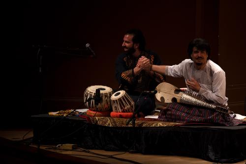 Two men seated on stage, holding hands.