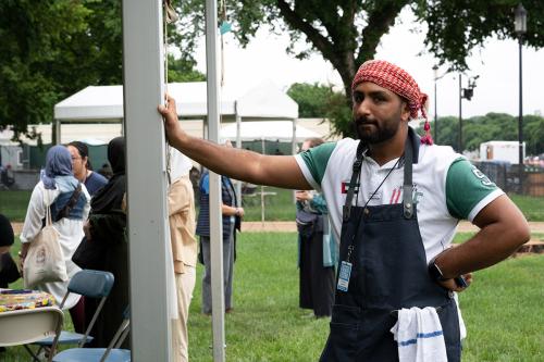 A man in chef's apron stands with one arm akimbo, resting against a tent pole.