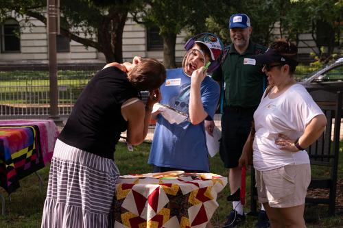 A young man holds a sewing circle up to his face, sticking out his tongue. A woman with her back to the camera does the same as a man and a woman look on, smiling.