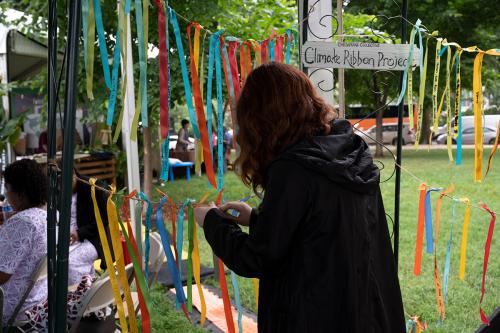 From behind, a person ties a ribbon to a line, adding onto four lines of many colored ribbons. A sign reads Climate Ribbon Project.