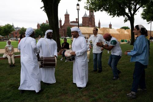 Three men in matching white shirts and headdresses perform on a wooden stage, two with drums and one with a trumpet-like reed instrument.