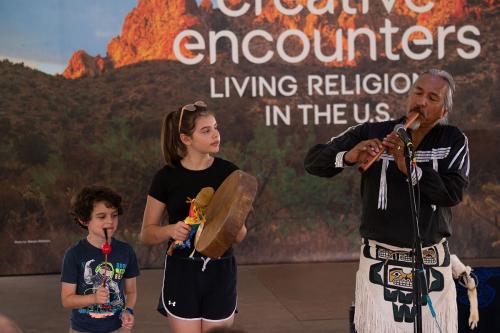 A Native American man plays a flute-like instrument into a microphone. To his right, a young girl plays a handheld drum-like instrument. To her right, a younger boy shakes a rattle-like instrument.