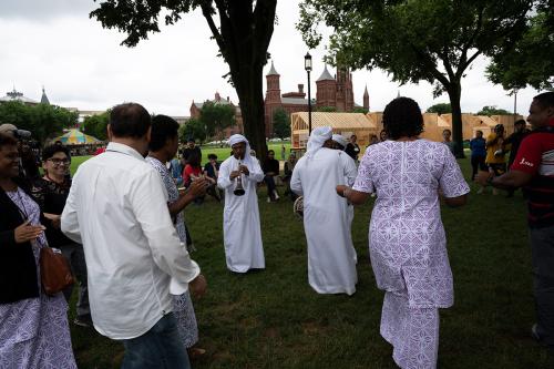 A small crowd of people dancing, including men in all white, one playing a shawn wind instrument, and women in purple printed dresses.