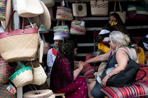 A seated woman in traditional Emirati dress applies a henna design to another seated woman’s outstretched hand. They’re seated on colorful, striped cushions in front of two other individuals in Emirati dress and a wall of hanging, woven baskets. 