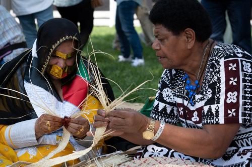 Two women, one in traditional Fijian dress and the other in traditional Emirati dress and covered by a Burqa, weave palm leaves together. They are sitting together in the grass.