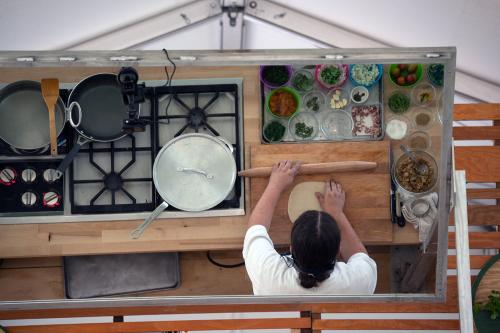 In a mirror from above, a man rolls out a circle of dough with a wooden pin. In front of him, various ingredients in bowls, and pans on the stove to his left.