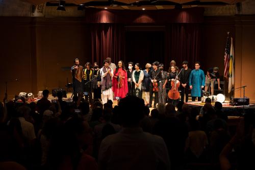 Performers on stage, standing together for a final bow, with audience giving standing ovation in the foreground.
