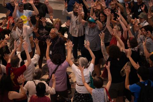 From above, a group of people gather in a circle, their hands raised in the air. At the center of the circle, a Balinese man chants.