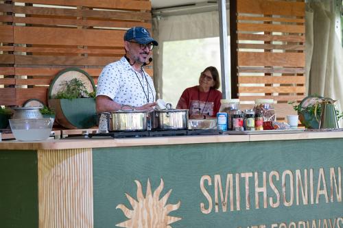 A man wearing a headset microphone smiles, working at a demonstration kitchen stove. His counter is full of pots and jarred ingredients.