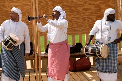 Three men in matching white shirts and headdresses perform on a wooden stage, two with drums and one with a trumpet-like reed instrument.