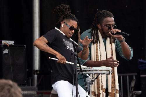 Two men performing on stage; on the left, a drummer with one stick in hand, one tucked under his chin, and one rapping into a microphone.