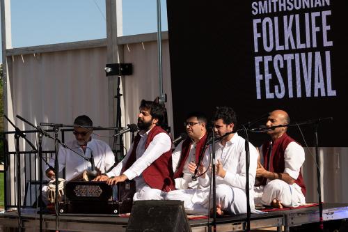 Five men performing on stage in matching white shirts and pants, some with red vest. Two in front play harmonium and tablas while others clap. Smithsonian Folklife Festival logo on the screen behind them.