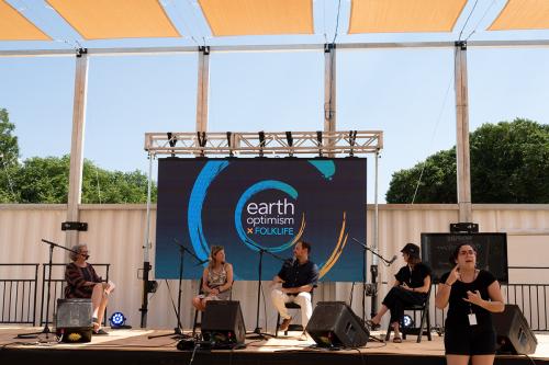 Four individuals sit on an elevated stage in front of a blue screen with the logo reading “Earth Optimism × Folklife Studio.”  A sign language interpreter stands in front of the stage. 