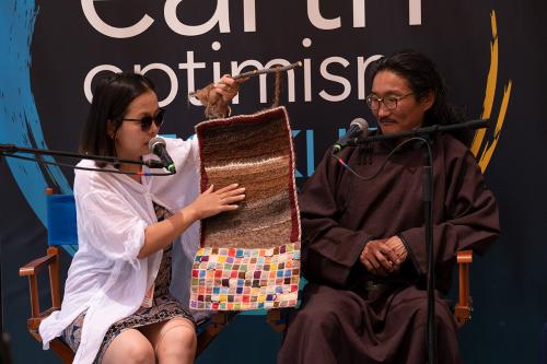 A man on woman sit on stage with microphones. The woman holds up and points to a small woven textile, with several striations of brown felt on top and squares of bright colors on the bottom.
