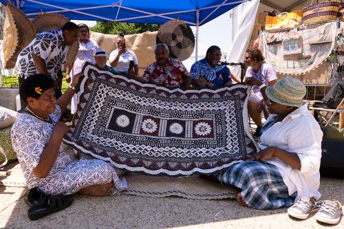 Two women sit on the ground, holding up a piece of artwork on barkcloth, printed with geometrical designs. A small crowd of people around them watch and cheer.