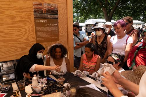A woman in a black head covering leads a workshop around a table, with bottles and jars of various liquids and flowers on the table. A crowd of people watch.