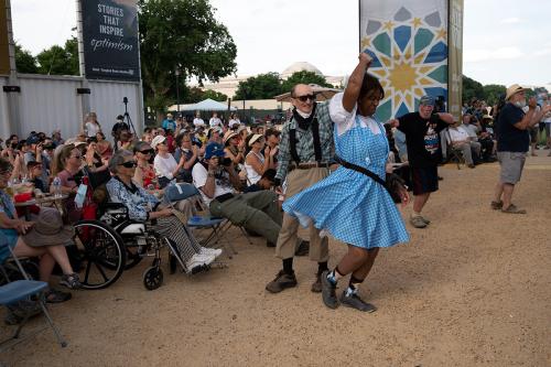 Between a crowd of people seated outdoors and a stage just out of frame, four people dance, including a man spinning a woman in a blue gingham dress.
