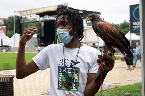 A man holds a large, brown bird in a raptor glove for protection. The bird and the man are looking to the left, while the man gesticulates.