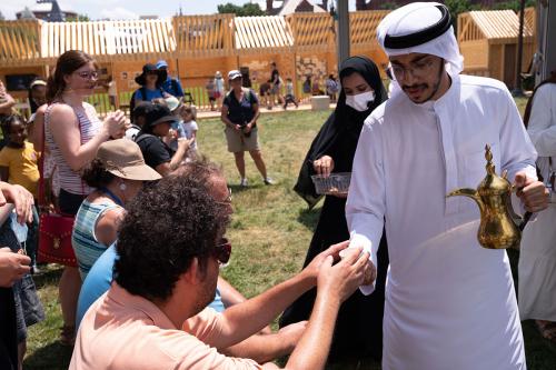 The man and a woman dressed in black hand out cups of coffee and dates to audience members seated and standing in the grass.