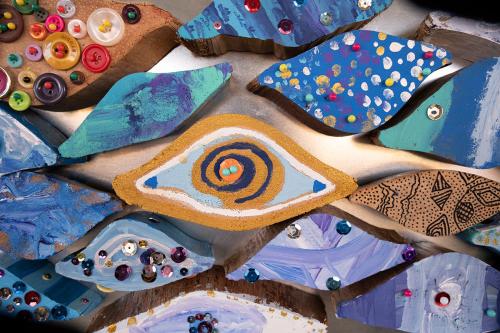 Close-up on several eye-shaped pieces of artwork arranged on a silver surface. They are each hand-painted, some with sequins and buttons glued on.