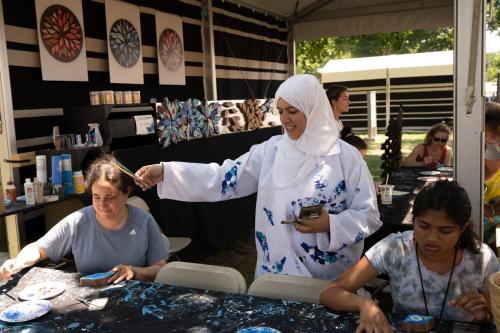 A woman in white dress and headscarf hands two paintbrushes across a table. The black tablecloth is splattered with blue paint. Two people sit at the table, working on painting eye-shaped pieces of palm.