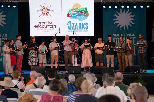 A chorus of fifteen singers stands in a horseshoe on the Ralph Rinzler Main Stage, one man standing slightly ahead to lead the audience in song.