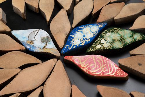 Close-up on eye-shaped pieces of palm. Most are unadorned in natural tan color, but four in the center are painted, one with a scene of a sailboat next to an island with a single palm tree, and three in blue, green, and red with gold Arabic calligraphy.