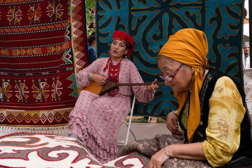 A woman sits in a chair playing a guitar-like Kazakh instrument. She is wearing a red patterned dress and a red circular hat with fringe on the sides. In the foreground, another woman wearing a yellow headwrap and a yellow shirt works on a felted quilt.