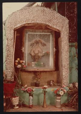 Small, three-tiered  altar holding a statue presumably of Our Lady of San Juan de los Lagos encased in glass. The lower two tiers have mismatched vases and candles with flowers. The altar has a frame of gray pebbles.
