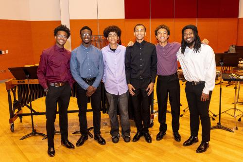 Six young people wearing dress shirts, slacks, and shoes pose arm in arm in front of a variety of musical instruments.