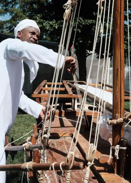 A man in traditional Emirati dress lifts the white sails of a small, model wooden boat.
