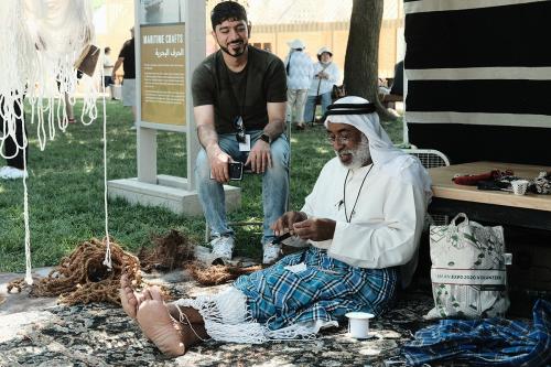 A man in white shirt and head covering sits on the ground, working with black yarn. Another man sits in a chair, smiling and watching him. Coils of rope hang around them and lay on the ground.