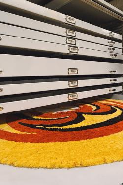 An open flat-flat drawer contains a red, yellow, and black feathered cape.