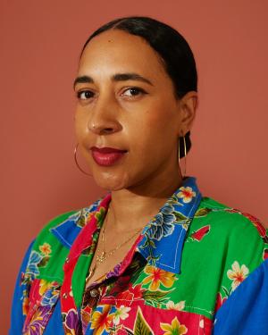 Headshot of Alexis Garcia, who has brown skin and dark hair tied back. She wears a bright floral collared blouse in blue, green, and pink.