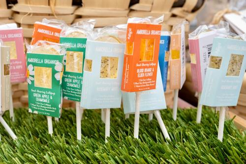 A group of lollipops in a display, each with a brightly colored label in green, orange, pink, or blue. The labels read Amborella Organics and various flavors: Blood Orange & Elder Flower, Green Apple & Chamomile, and Rosemary Mint.