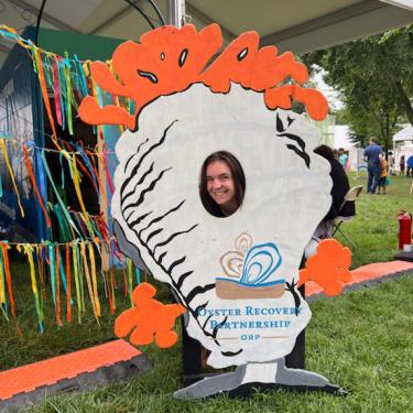 Young woman smiles, her face in the cutout of a photo booth prop depicting a white oyster shell with bright orange appendages. Text on it reads Oyster Recovery Partnership.