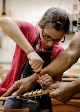 A girl in a red T-shirt and dark apron uses a screwdriver on the headstock of an acoustic guitar. An adult, out of frame, braces the instrument for her.