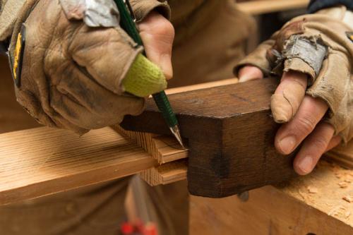 Close-up on a pair of fingerless-gloved hands marking a piece of lumber with a pencil. Both point fingers are bandaged.