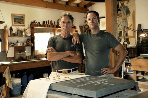 Two men posing together inside of a stone carver's workshop, one with crossed arms and one with an arm resting on the other's shoulder.