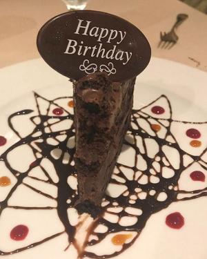 A small slice of chocolate cake on a white plate drizzles with chocolate syrup; the cake slice is topped with a chocolate decorative oval that says Happy Birthday. 